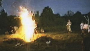about 15 feet high, shaped like a winged being, appearing on the left, surrounded by night trees and a field, and a figure on the right, facing the fire